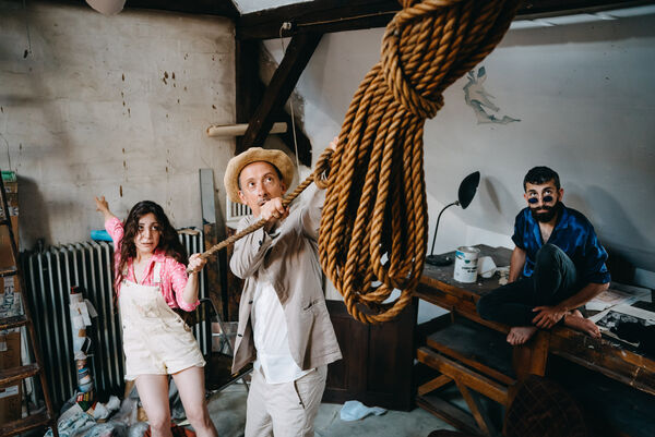 Eine Frau mit langen dunklen Haaren (kurze helle Latzhose und rosafarbige Bluse mit weißen Punkten) und ein Mann mit Sonnenhut (und hellem Leinenanzug) ziehen an einem Tau. Ein im Hintergrund hockender Mann mit dunklen kurzen Haaren und schwarzem Vollbart beobachtet das Tauende über seine Sonnenbrille hinweg. 