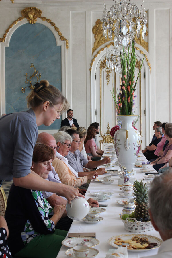 Veranstaltung „Teegenuss beim König“ in den Neuen Kammern, Menschen an gedeckter Tafel, eine Frau schenkt Tee ein